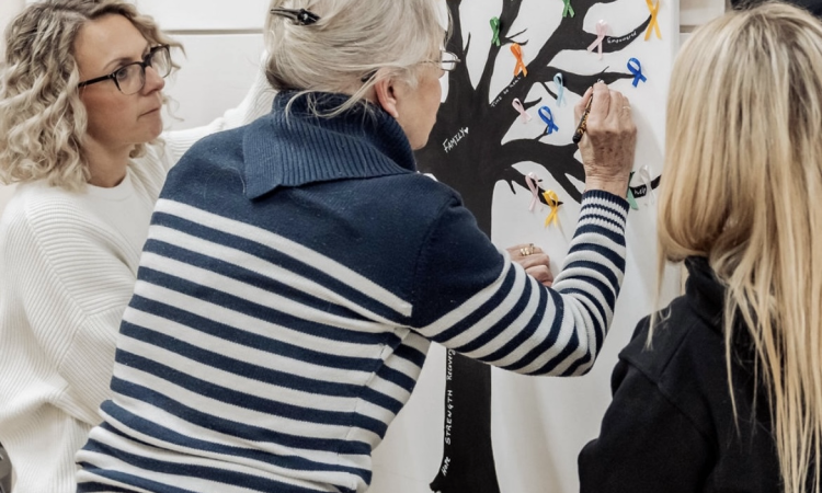 A woman adds finishing touches to a painting of a tree, helped and watched by two others.